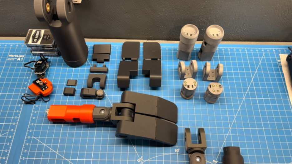 Wall-E arm components laid out on a cutting mat, including black 3D-printed shoulder and forearm parts, gray MJF nylon pieces, the orange glass-fiber wrist section, and micro servos