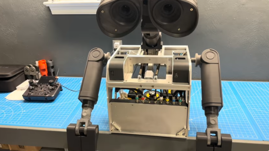 Life-size unpainted Wall-E robot with binocular head and articulated arms standing on a blue cutting mat in the workshop